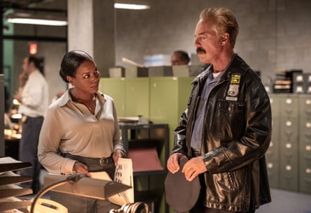 A policeman in a black leather jacket talks to  woman standing over a desk.