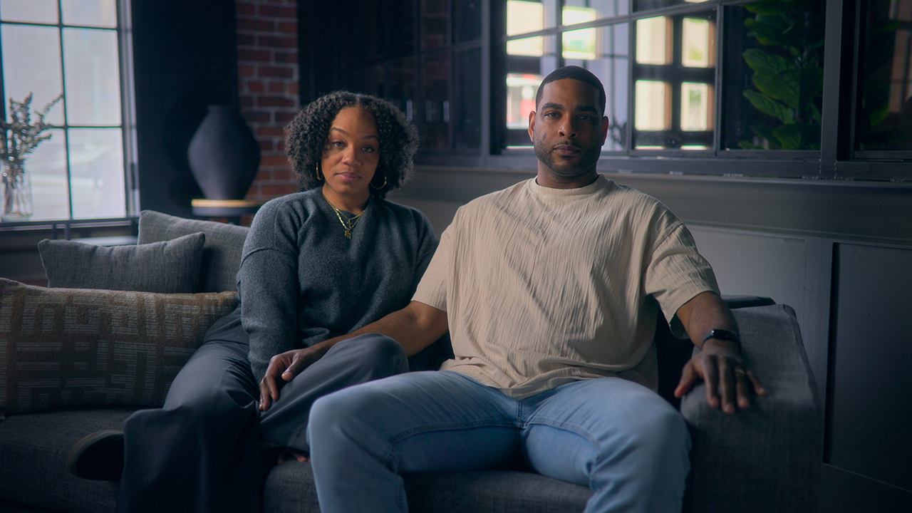 &Quot;Two People Sit Closely Together On A Couch In A Modern Living Room With A Large Window, Exposed Brick Wall, And Soft Natural Lighting, Both Looking Toward The Camera.&Quot;