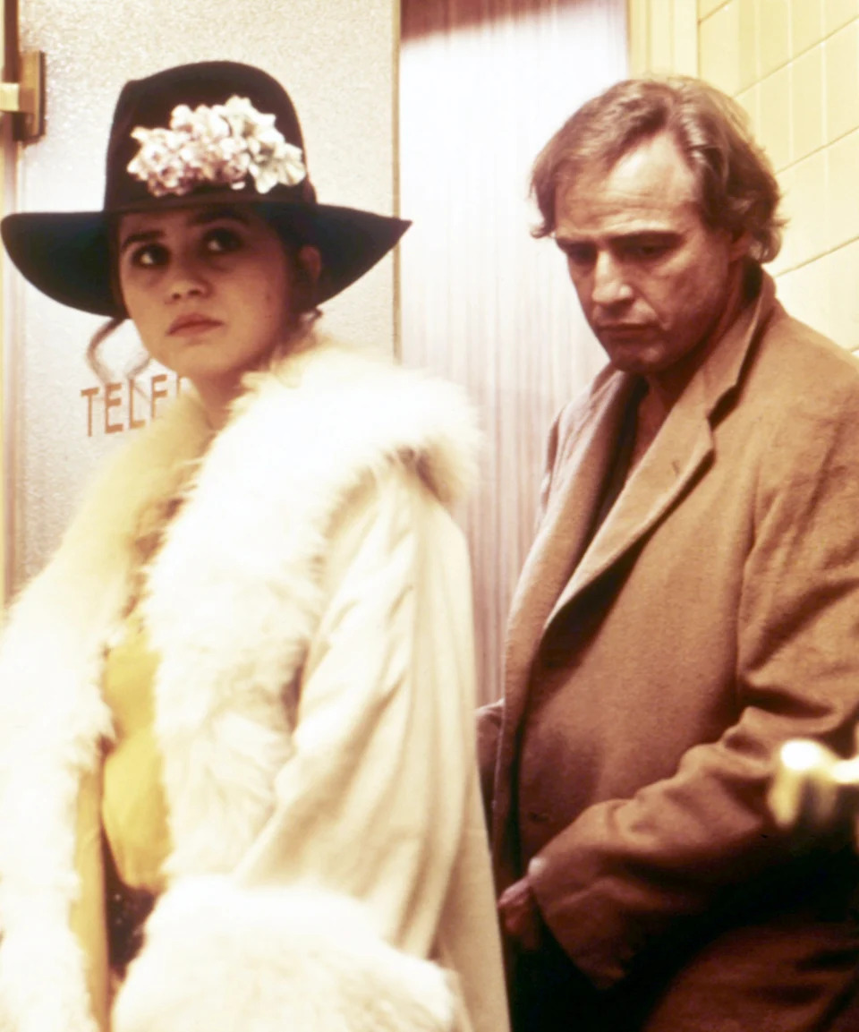 Marlon in a coat stands behind Maria in a hat adorned with flowers and a fur-trimmed coat, in a scene suggesting tension or drama