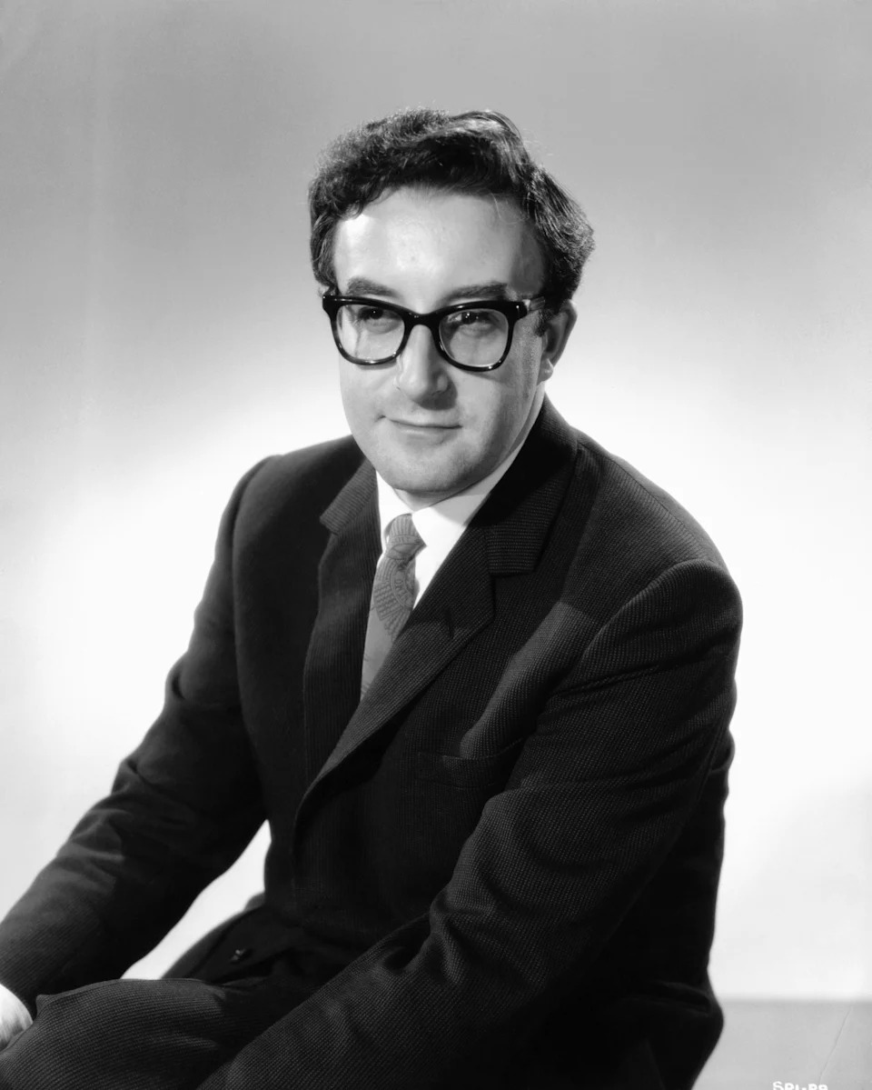 Peter Sellers in a suit and tie sits facing the camera, wearing glasses and with a neutral expression