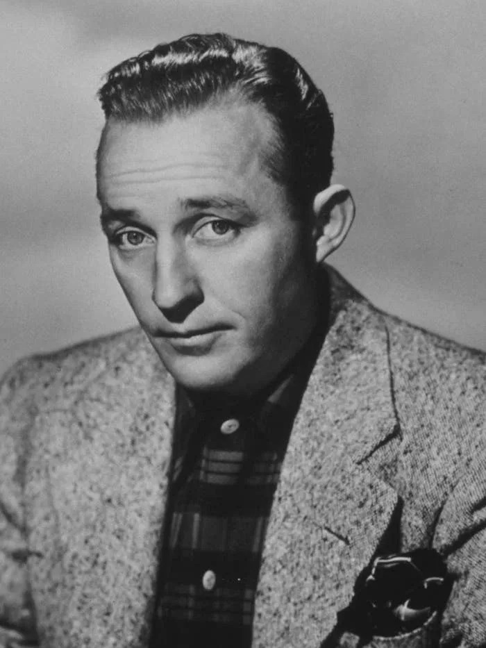 Bing Crosby in a 1940s-era tweed suit and plaid shirt, seated with papers, looking thoughtfully at the camera