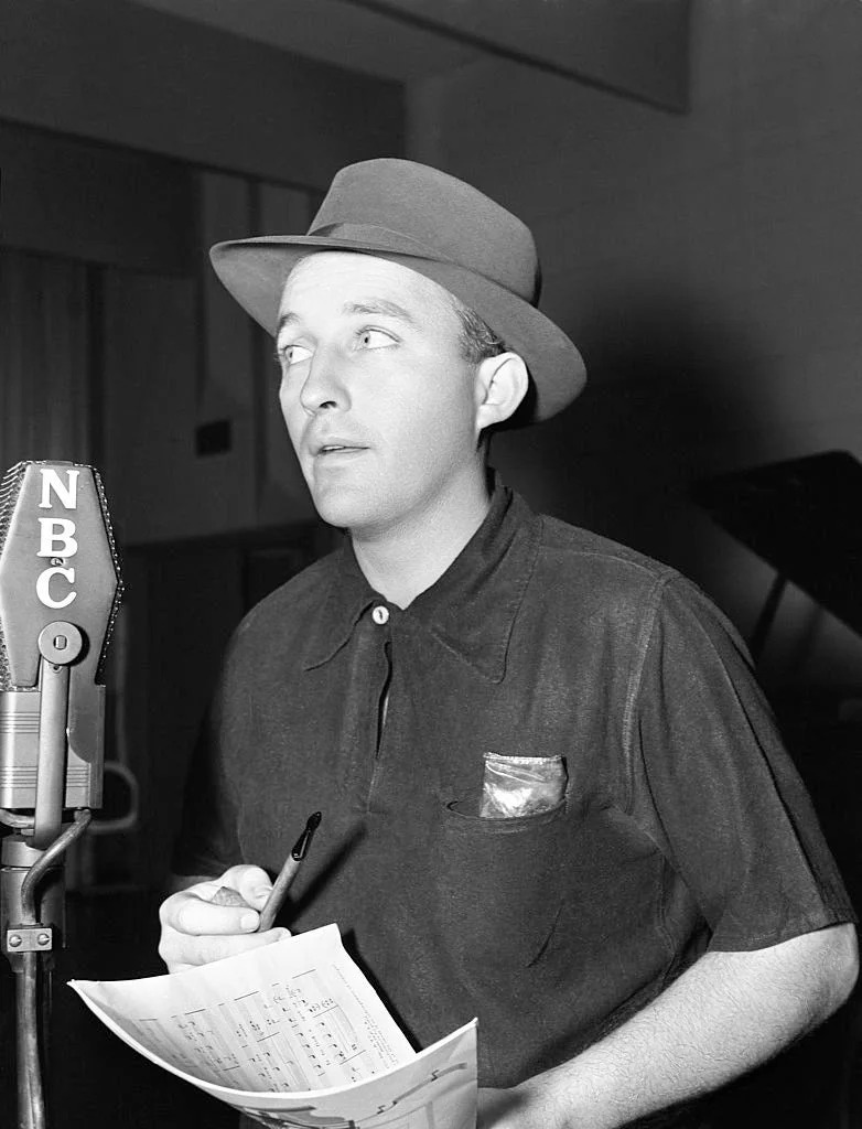 Person in vintage-style shirt and hat stands at an NBC microphone holding a pen and sheet music, appearing focused