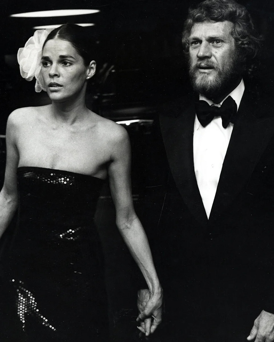 Steve in a tuxedo and Ali in a strapless sequin gown with a large headpiece walk hand in hand at a formal event