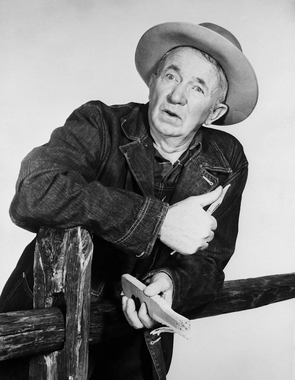 Walter Brennan in denim jacket and cowboy hat leans on a wooden fence, holding a piece of wood, with an expressive facial expression